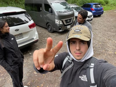 This image depicts an outdoor scene on a dirt and gravel path, identified as occurring in the city of San Rafael, Mexico, where several individuals and vehicles are present.

In the foreground, a young adult male with dark hair, wearing a beige "VAIL 1966 COLORADO" baseball cap over a light grey hoodie and a dark grey "THE NORTH FACE" long-sleeved shirt with a grey backpack, takes a selfie. His right hand is extended, displaying a two-finger peace sign towards the camera. He maintains a neutral expression.

To the left, a young adult female with dark hair, wearing a black North Face jacket, black pants, and partially visible white shoes, stands facing towards the right with a slight smile. Her hands are in her pockets.

Behind the foreground male, a second young adult male with dark curly hair and a grey long-sleeved shirt is visible. He is smiling and looking towards the female on the left.

Parked behind the individuals are a line of vehicles on the dirt path. From left to right:
1.  A white Kia SUV (likely a Carnival or Sorento) with an "ESTADO DE MEXICO" license plate (018-BAR).
2.  A grey Nissan NV350 Urvan passenger van with a roof rack and a partially visible license plate (DDF-213-B), facing away from the camera.
3.  A white sedan or hatchback (possibly a Renault model) with a partially visible license plate (21-WXF-8), also facing away.
4.  A blue hatchback (possibly a Chevrolet model) is parked farthest in the background on the right, facing away from the camera.

The environment consists of dense green foliage and trees in the background, suggesting a natural or wooded area. The ground is composed of compacted dirt and small stones, with scattered leaves. The lighting indicates daylight, possibly overcast.