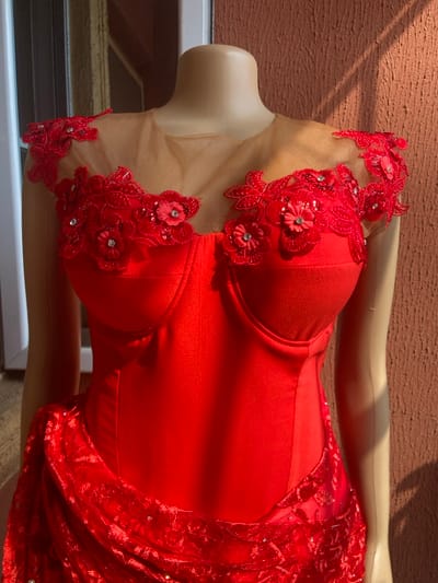 The scene depicts a red formal gown displayed on a light-skinned, armless female mannequin, set outdoors against a building in Oba-Aboji, Nigeria. The dress features a sheer, light brown illusion neckline adorned with intricate red floral lace appliques, some accented with small red fabric flowers and central clear rhinestones, extending across the shoulders and upper chest. The bodice is structured with visible bust cups and vertical seam lines, indicating a form-fitting design. The lower section of the dress, partially visible at the bottom left, consists of draped, ruched red fabric with a patterned lace overlay, which appears to contain subtle reflective elements. The mannequin and dress are positioned against a textured, light brown wall, possibly stucco or concrete. A vertical white architectural element, likely part of a door or window frame, is visible on the left. Strong natural sunlight illuminates the scene, casting distinct shadows on the wall behind the mannequin.