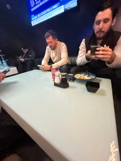 The image displays an indoor scene within a dimly lit commercial establishment, likely a lounge or cafe, situated in Illkirch-Graffenstaden, France. Four adult males are visible, primarily focused on their personal electronic devices.
In the foreground, a large, light-colored, reflective rectangular table is present. Partially visible on the left side of the table are hands holding a smartphone. Further right on the table, a red beverage can stands next to a black-based salt and pepper shaker set. Adjacent to these are a blue bowl containing partially consumed pasta with a fork, and a small, empty black square bowl.
Two males are seated at the table in the mid-ground. The male on the left, characterized by short brown hair and a beard, wears a light-colored long-sleeved top and is actively viewing a smartphone held in his hands. To his right, the second male has dark hair and a beard, wears a white long-sleeved shirt beneath a dark vest displaying "R.1" text, and is also engaged with a smartphone in his hands.
In the background, on the far left, a third male is dimly perceptible, wearing a dark cap and jacket. He holds a hookah pipe to his mouth while simultaneously looking at a smartphone. Two light-blue shisha pipes are positioned on a separate white table to his left. A fourth, unidentifiable figure is seated further back, also appearing to use a phone. A large television screen is mounted high on the dark wall, displaying French news captions, including "DOSSIER : QUELS RECOURS POUR LA FRANCE ?". The seating throughout the establishment consists of dark, upholstered couches or benches. The overall atmosphere suggests a casual social gathering where individuals are predominantly engaged with their smartphones.