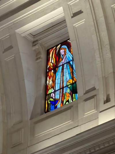 A vertical rectangular stained glass window is centrally displayed within a deep, light-colored architectural recess. The window is located within a religious structure at the Sanctuary of Fátima, Fátima, Portugal, consistent with the interior of the Basilica of Our Lady of the Rosary. The surrounding architectural elements are composed of uniform light-toned stone or plaster, featuring prominent cornices, a decorative corbel on the upper left, and vertical fluted details on the right side of the window opening.
The stained glass depicts a religious scene. A tall, veiled female figure, rendered in shades of light blue and white, dominates the upper and central portion, with her hands appearing pressed together. Behind this figure, predominantly to the left, are abstract forms in red, orange, and yellow, suggestive of flames or emanating light. In the lower section of the window, three smaller figures are portrayed, appearing as children. Two figures on the lower right, in green and orange attire, are depicted in a bent posture, suggesting kneeling or bowing, with their gazes directed upwards. A third figure on the lower left, dressed in blue and green, also looks upwards towards the central figure. Dark leading defines the figures and segmented glass shapes, which consist of a vibrant palette including blues, whites, reds, oranges, yellows, and greens.