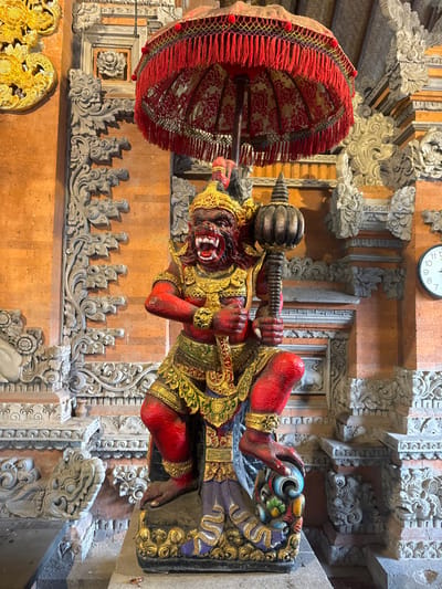 The scene depicts a vividly colored, ornate statue positioned indoors against a backdrop of traditional Balinese architecture in Batuan, Indonesia. The central subject is a large, crimson-skinned figure, likely a guardian deity, with a ferocious expression, bared fangs, and red eyes. This figure wears a golden crown, intricate golden armor, and jewelry on its arms, chest, waist, and legs, with additional green and purple accents. It holds a dark, mace-like implement in its raised right hand and is depicted in a crouching stance, with its left foot resting on a multi-colored, swirling decorative base. Directly above the statue, a ceremonial red umbrella (payung) with red fringe and a patterned fabric is positioned. The background features terracotta-colored brick walls extensively adorned with grey stone carvings depicting complex floral and mythological motifs. A circular white clock with black numerals is partially visible on the right wall, and a golden ornate carving is on the far left wall. The floor is dark and textured. No individuals are present.