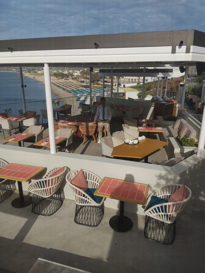 The image shows an elevated outdoor dining area, likely part of a restaurant or bar. The location is Pyli, Greece, as suggested by the beach and ocean view visible through the structure's clear walls. Tables are arranged with wicker-style chairs and red tabletop details. Some seating areas include comfortable pillows. A person dressed in light-colored clothing with a blue tie is visible standing between the tables, possibly a waiter or manager. The area appears to be well-maintained, with a modern architectural design that incorporates both open-air and enclosed spaces. The sky is blue with some clouds, indicating a pleasant weather condition.
