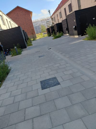 The image depicts a modern residential street with newly constructed brick houses on the right and dark wooden-clad annexes, possibly storage units or garages, on the left. The street is paved with light grey rectangular cobblestones, and a narrow, curved drainage channel runs down the center of the pathway. In the foreground, a square manhole cover is visible on the cobblestones. To the left, near the wooden annexes, there is some green vegetation and a blue and orange child's stroller. The sky is clear with a few wispy clouds, and the sun casts shadows, indicating it is likely daytime. In the background, scaffolding is visible around a building, suggesting ongoing construction. The architectural style suggests a contemporary development in a suburban area. This scene is located in Taastrup, Denmark, specifically at the residential street of Grøndalsparken.