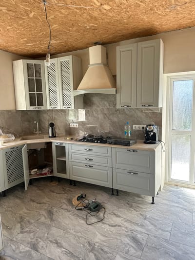 The image depicts a kitchen area that appears to be under renovation or recently completed. The scene is set indoors, with light streaming in from a large window at the top, suggesting daytime. The floor is covered in large, grey marble-patterned tiles.
The main subjects are kitchen cabinets. On the left, there are light-colored cabinets with decorative lattice fronts and glass-paneled doors. Above these, a beige range hood is visible. On the right, more light-colored cabinets are present, including upper cabinets with decorative fronts and lower cabinets with drawers and open shelving. Some of these cabinets are still in the process of being installed, as evidenced by exposed legs and plumbing beneath one of the countertops.
On the countertop, a black coffee machine and a bottle of water are visible, plugged into a white electrical outlet. A stovetop is also installed. Tools are present on the floor, including a jigsaw and a sanding tool, indicating ongoing work. A lightbulb hangs from a wire attached to a plywood-covered wall on the far left, also suggesting construction or renovation. The overall atmosphere is one of a functional space being prepared or updated. There is no visible text in the image.