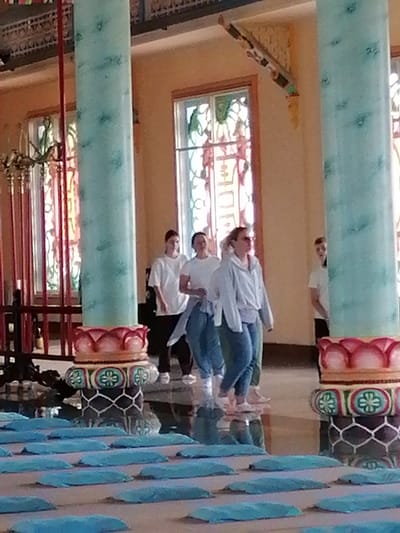 In Lộc Quý, Vietnam, inside a building with ornate pillars and stained-glass windows, a group of people, primarily young women, are walking. The women are dressed casually in t-shirts, sweatshirts, and jeans. The floor is highly reflective, mirroring the pillars and the people. In the foreground, a series of light blue cushions are neatly arranged. The overall atmosphere suggests a cultural or religious setting, possibly a temple or meditation center.