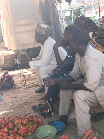 The image depicts an outdoor market scene in Kawaji, Nigeria. Three adult males are seated side-by-side on a low surface, likely the ground. The leftmost man, appearing older, wears a white striped long-sleeved garment and a patterned white and blue kufi cap. He holds green currency in his left hand, and his feet are clad in black shoes. The central man wears a dark short-sleeved t-shirt with a light graphic and dark trousers. His right hand is positioned near his chin, and he holds a string of black prayer beads (tasbih) with both hands. Dark flip-flops are on the ground near his bare feet. The rightmost man wears a light beige long-sleeved tunic and matching trousers. He is also barefoot, with dark flip-flops beside him.
In the foreground, a large pile of red and green peppers, along with some small red tomatoes, rests on a light-colored mat. A light green plastic bowl and a blue plastic bowl are positioned near the produce. To the left, a bowl of red onions is visible, and a black string of beads is displayed on a small dark stand. In the midground, behind the seated men, makeshift market structures are present. The background shows blurred market activity, including a green motorized tricycle (keke napep) and a stall with a pile of brown, spherical items, possibly coconuts or yams. The scene is illuminated by bright daylight, indicating an open-air environment with dusty ground.