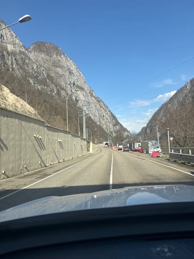 The image captures a mountain road in Monastyr', Russia, on a clear, sunny day, viewed from inside a vehicle looking forward. The road is asphalt, two-laned, with white solid and broken lane markings, and appears to be ascending or winding through a valley. Steep, rocky mountainsides, sparsely vegetated with leafless trees, flank both sides of the road under a bright blue sky, which features a faint aircraft contrail.
On the left side of the road, a long, segmented concrete retaining wall parallels the road. Multiple streetlights on tall poles are positioned along this wall. Further down the road, a dark tunnel entrance is visible on the left. Shadows are cast by the wall and roadside objects, indicating direct sunlight from the right.
Ahead, near the tunnel entrance, road construction or maintenance activity is evident. A white truck, possibly equipped with a crane, is positioned in the right lane, followed by a dark sedan. On the right shoulder, a temporary site features various informal structures, possibly sheds or containers. A red vehicle is parked near these structures, and a significantly damaged white vehicle, possibly a car with "AMG" visible on its side, rests precariously against some of the temporary structures and red barricades. A metal guardrail runs along the right edge of the road, and a white triangular "give way" sign with a red border is visible. A distant, snow-capped mountain peak can be seen through the valley opening. No individuals are distinctly visible.