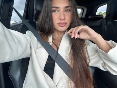 A woman is shown in an upside-down orientation inside a car with black leather seats. She wears a white blazer and a black top. She has brown hair and is wearing a seatbelt. A view through the car window shows trees and buildings, indicating an urban or suburban environment. The image was taken in North Miami, United States of America.