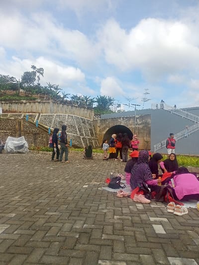 This image depicts an outdoor gathering of people in Pangalengan, Indonesia, under a partly cloudy sky during daylight hours. The scene is set on a large paved area made of interlocking bricks, with various concrete structures and natural elements in the background.
In the foreground to the right, a group of women, many wearing hijabs, are seated on the pavement. They appear to be resting or having a picnic, with shoes, bags, and what look like drinks scattered around them. Several individuals are standing and conversing near a concrete archway or tunnel entrance in the mid-ground. Two people walk across the paved area on the left. Further back, a person in a red jacket and backpack is visible on a set of white stairs leading up a gray building.
The background features terraced concrete retaining walls supporting a hillside covered with trees and lush greenery. Light poles with what appear to be solar panels are also visible. On the far left, a large object covered with a white or gray tarp rests on the ground. The lighting suggests late morning or early afternoon, with soft shadows and a mix of blue sky and white clouds indicating pleasant weather. People seem relaxed and engaged in social interaction. No discernible text is visible.