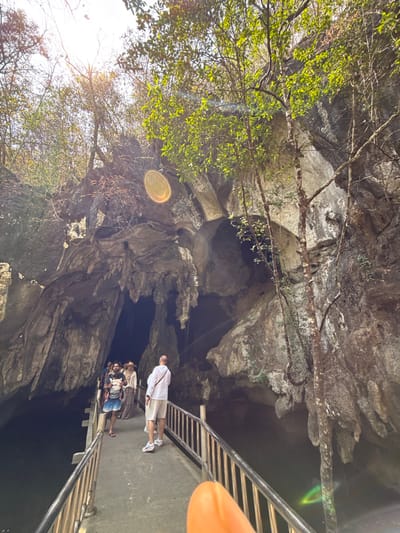 A group of people stand on a walkway leading into a large cave. The cave entrance is surrounded by rock formations and trees. Sunlight streams in from the left, creating lens flares. The walkway is elevated and has metal railings. A person in a white shirt and tan shorts stands at the front of the group.