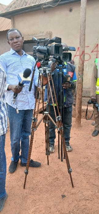 The image shows a media scene in Rigasa, Nigeria, on an unpaved reddish-brown dirt ground. Three male individuals are present with professional video and photographic equipment. In the foreground left, a man wearing a light blue and white striped shirt and blue jeans holds a black microphone with a white cube-shaped wind screen, looking towards the viewer. Central to the frame, a large professional video camera is mounted on a brown, three-legged tripod. Partially visible behind this camera, a second man, likely a cameraman, wears a dark vest and appears to be operating the video equipment. To the right, a third man, wearing a light-colored vest, holds a black handheld camera. The background features a light brown building with a corrugated metal roof, displaying red graffiti, including the partial characters "A54," on its wall. A vertical wooden pole stands to the right of the video camera.