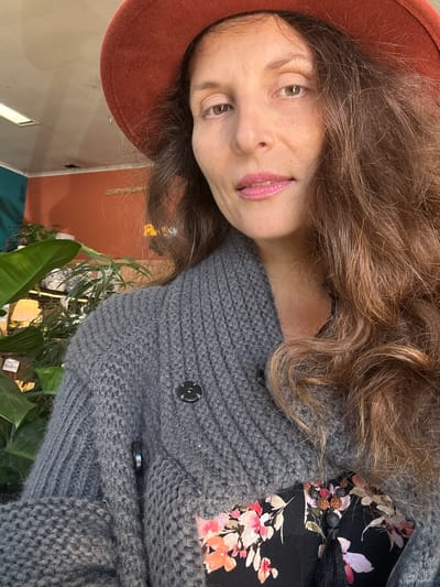 A woman wearing a rust-colored hat and a grey cardigan is pictured in a close-up shot. She has long, wavy brown hair and is looking towards the camera with her lips slightly parted. The background appears to be an indoor setting with plants visible. The location is specified as the city of Bellingen, Australia.