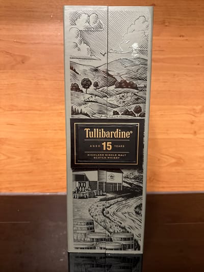 The image shows the side of a Tullibardine 15 Years Old Highland Single Malt Scotch Whisky box. The box has a detailed illustration of a rural landscape with rolling hills, trees, livestock, and a building that appears to be a distillery. There is also an illustration of barrels. The text on the box clearly states "Tullibardine", "AGED 15 YEARS", and "HIGHLAND SINGLE MALT SCOTCH WHISKY". The box is positioned vertically against a wooden background, likely a table or shelf. No people or any specific location in Silvolde, Netherlands are visible.