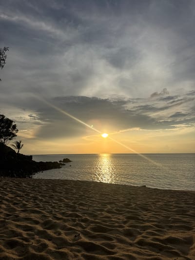 The image depicts a coastal scene in Baan Wang Ta Kien, Thailand, at sunset. The sun is positioned near the horizon, emitting bright rays that reflect off the water. The sky features a mix of clouds, some dark and others lighter, indicative of variable weather conditions. The water appears calm with visible sun glint. The foreground includes a sandy beach with textured patterns, and vegetation (trees, possibly palms) along the coastline. The orientation is likely vertical.