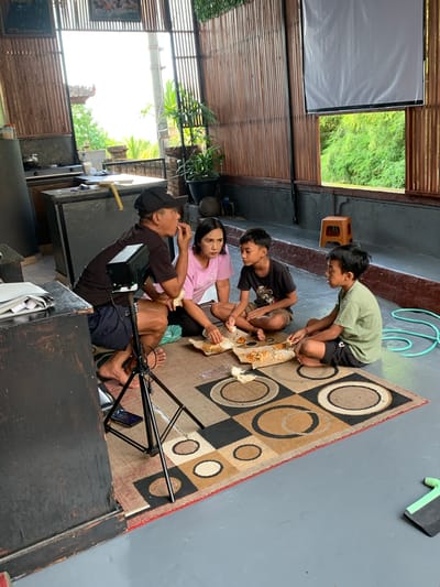 In Sukasada, Indonesia, a family is gathered on a mat on the floor, eating a meal. A man wearing a black t-shirt and a cap is eating and appears to be talking to the woman next to him, who is wearing a pink t-shirt. Two boys, one in a black t-shirt and the other in a green t-shirt, are also eating. They are all sitting on a patterned mat, with their food served on paper plates. The environment appears to be an open-air covered area with bamboo walls and a grey floor. There is a tripod with a small device on it, a wooden table, and some plants visible. A projection screen is mounted on the bamboo wall.