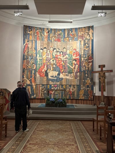 The image depicts the interior of a church, likely in Ribeauvillé, France, featuring a large, colorful mural that spans the rear wall. The mural is composed of wooden panels and depicts numerous figures in a narrative style, possibly religious in nature. In the foreground, two men are standing, facing the mural. One man, on the left, is wearing a dark jacket and jeans, and the other man is partially visible behind him. To the right of the mural stands a wooden crucifix with a figure of Jesus. The church interior also includes wooden chairs, a patterned carpet on the floor, and a raised altar area with steps. The lighting appears to be artificial, with spotlights visible on the ceiling. The overall atmosphere is one of reverence and contemplation.