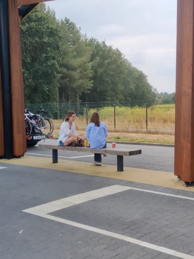 Two women are sitting on a wooden bench in an outdoor seating area next to a parking lot. The woman on the left is wearing a white shirt and blue shorts and is looking to her right. The woman on the right is wearing a blue shirt and dark pants and is looking away from the camera. A bicycle rack with two bicycles is attached to a black car on the left side of the image. The background consists of trees and a fence. The scene is located in the city of Maarheeze, Netherlands.