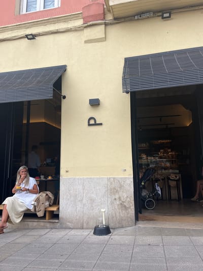 This image depicts the exterior of a commercial establishment, identified as a cafe or bakery, located in Santander, Spain. The building features a light yellow-cream facade with a light grey marble-tiled lower section. Two open doorways, each with black-framed glass doors and topped by a black slatted awning, provide entry. Between the doorways, a stylized black 'p' logo is affixed to the wall, positioned below a small black rectangular wall-mounted light. Above, the building transitions to a reddish-pink section with a white-framed window, equipped with bird deterrent spikes along its lower edge. Security or floodlights are also visible on the upper facade.
On the left side, a blonde woman, dressed in a white short-sleeved top and light beige wide-leg pants, is seated on a wooden bench positioned just inside the entrance. She holds a small yellow cup on a saucer in her left hand and brings her right hand to her mouth, appearing to eat. A light brown jacket is draped on the bench beside her. Further inside this entrance, a man in a light blue shirt is visible behind a counter.
The right doorway offers a view into the establishment, revealing a display case filled with baked goods or pastries. A black baby stroller is parked near this entrance. On the grey square-paved sidewalk directly in front of this entrance, a black pet food or water bowl, featuring a prominent white bone-shaped handle, is positioned on the ground. The overall scene suggests a functional urban environment.