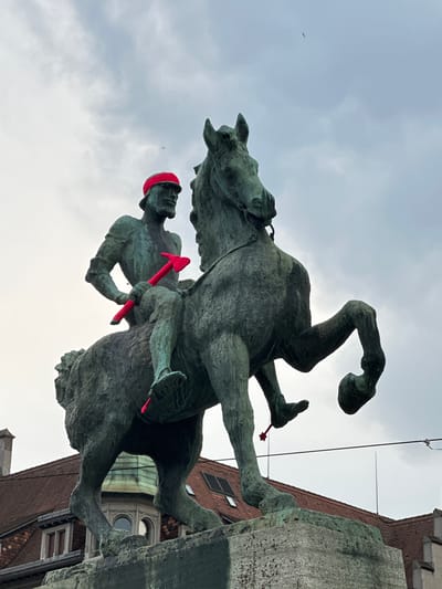 The image shows a bronze equestrian statue in Zurich, Switzerland, adorned with bright red knitted accessories. The statue depicts a man on horseback, wearing a red bandana on his head and holding a red, knitted axe. A similar red knitted item is attached to the horse's hind leg. The statue is positioned in front of a cloudy sky, with the rooftops of buildings visible in the background. The architecture of the buildings suggests a European setting, consistent with Zurich.