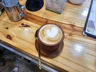 A cup of latte art sits on a wooden table, accompanied by a golden spoon. Beside the cup, there's a stainless steel pitcher and a white carton. A black object is partially visible near the pitcher, while a menu or document with a black border lies to the right. The background features a glimpse of flooring and possibly the base of a cabinet or counter.