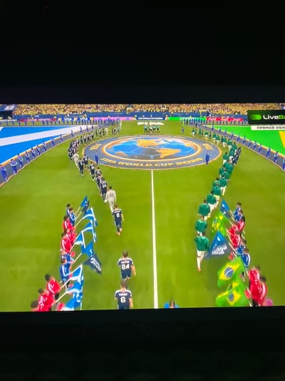 The image depicts a football (soccer) stadium during what appears to be an opening ceremony. Two lines of individuals, likely football players in uniform, are walking onto the field from opposite sides. One line of players is wearing predominantly green kits, while the other is wearing white and black kits with some red accents. Several individuals are carrying flags. On the left side, there are individuals holding blue flags with white designs, and on the right, there are individuals holding green and yellow Brazilian flags. A large circular logo with "FIFA WORLD CUP 2026" is visible on the field in the center. The spectators in the background are wearing yellow attire and are seated in the stands. The scene is well-lit, suggesting it is either daytime or a stadium with extensive lighting. There is no indication of the city of Slobozia-Dușca, Moldova, in the image.
