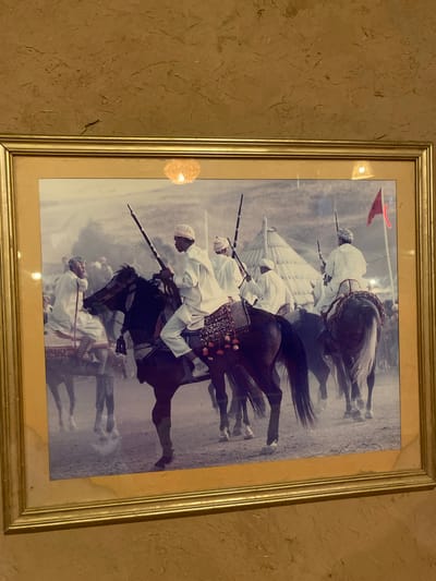 A framed, horizontal photograph depicts a group of men on horseback in what appears to be a desert or sandy environment. The men are dressed in traditional white robes and head coverings, and many are carrying long rifles or spears. They are riding dark-colored horses, some of which are adorned with decorative saddle blankets. In the background, there are tents or large conical structures, and a red flag is visible on a pole. The overall scene suggests a ceremonial procession or gathering. The location is specified as Agafay, Morocco, a desert region known for its arid landscapes and traditional Berber culture.
