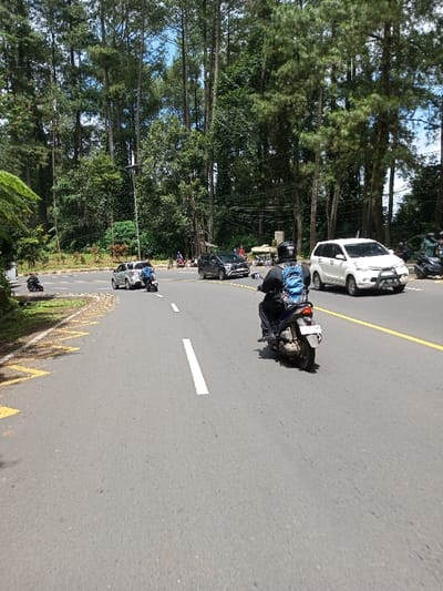 The scene depicts a sunny day on a multi-lane asphalt road in the city of Boommlog, Bhutan, surrounded by dense, tall pine-like trees. In the foreground, a person on a dark-colored scooter, wearing a black helmet and a blue backpack, travels away from the viewer on the right side of the road. The scooter has a rear license plate. The road features white lane markings and yellow solid lines, with a junction or turn-off to the left marked by yellow zigzag curb lines. At this junction, a silver sedan is either turning or waiting, with another scooter nearby. On the opposite side of the main road, a white SUV/MPV is driving towards the viewer. Further in the background, a dark SUV is visible, along with several other motorcycles and individuals. Utility poles are present amidst the trees.