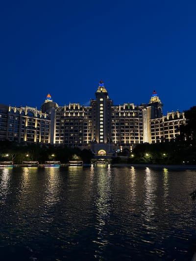 The image depicts a multi-story building with ornate architectural details and numerous windows, illuminated at night. The building reflects in a body of water, likely a river or lake, creating a mirrored effect of the lights. The sky is a dark blue. The photo suggests the location is Xiangzhou, China, as it matches architectural styles found in that region.