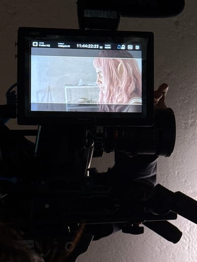 The image depicts a close-up view of a professional video camera's external monitor, showing live or recorded footage, situated in El Paraíso, Mexico. The monitor, displaying "HDR," "ProRes HQ" codec, and "1080p23.98" format at timestamp "11:44:22:22," features a profile shot of a female subject. This subject has long, wavy pink hair and prominent, pointed prosthetic elf ears. She is looking towards the left side of the frame and appears to be wearing a light-colored top. The background within the monitor's footage is an indistinct indoor setting with light walls.

The camera rig itself is largely silhouetted in the foreground against a textured, light-colored wall. A large camera lens extends from below the monitor to the right. A microphone is mounted atop the camera, above the monitor. A human hand, also silhouetted, is visible on the right side, appearing to hold or adjust part of the camera assembly. The immediate environment of the photograph is dimly lit, with a brighter, localized light source illuminating the background wall to the right of the camera.