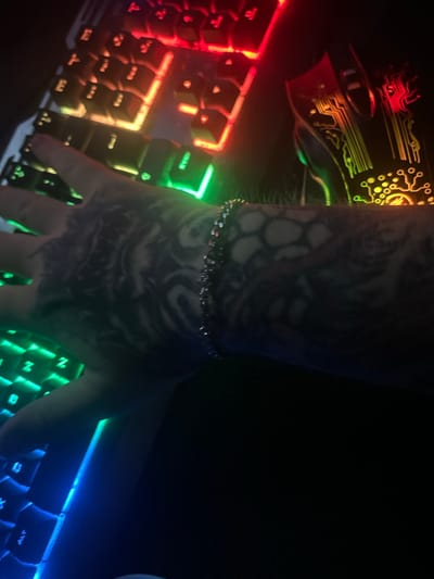 The image shows a close-up view of a person's tattooed arm resting on a keyboard with RGB lighting. The keyboard keys are illuminated with a gradient of red, orange, yellow, green, and blue light. To the right of the keyboard, a gaming mouse with a circuit board design and similar RGB lighting is partially visible. The person's arm features intricate black tattoos. The scene appears to be indoors in a dimly lit environment, possibly a gaming setup or computer desk. There is no information in the image to suggest a location in Gelsenkirchen, Germany.