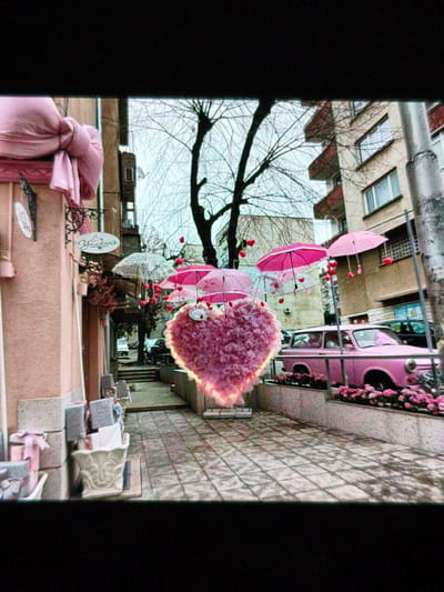 The image captures a street scene in Devin, Bulgaria, decorated for Valentine's Day. A large, pink heart-shaped decoration with a glowing aura hangs suspended in the air. Pink umbrellas with dangling red hearts are also suspended. A pink Trabant car sits behind a concrete planter box filled with pink flowers. Buildings line the street, and a tree with bare branches is visible between the decorations. Paved tiles cover the street. A shop or cafe on the left has pink decorations and a sign in Cyrillic. There are no people clearly visible in the image.