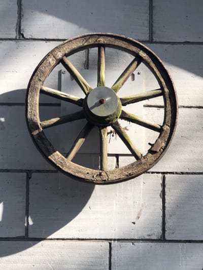An antique, weathered wooden wagon wheel is centrally mounted on a light grey block wall in Сапоцкінскі сельскі Савет, Belarus. The wheel features a central metallic hub, from which ten wooden spokes radiate to a wooden rim. The wood of the wheel shows significant signs of age, including rough texture, cracks, and patches of green moss, particularly around the hub and along the spokes. Remnants of a metal band or fasteners are visible on the upper right portion of the rim. The wheel is affixed to a rectangular, light-colored panel, which is secured to the wall by at least two visible fasteners. The wall is constructed from large, rectangular light grey blocks with visible mortar lines. Strong sunlight from the upper right casts a distinct, crescent-shaped shadow of the wheel onto the wall to its left and below, creating illuminated and shadowed areas on the block surface. No individuals are present in the scene.