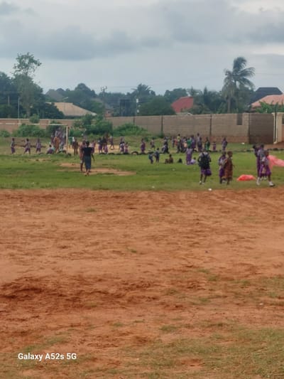 In Umudioka, Nigeria, a group of children are engaged in an outdoor activity on a dirt and grass field. The scene appears to be a school or community gathering, with numerous children scattered across the green grass area. Some children are in motion, while others are gathered in small groups. In the background, a brick wall and fencing delineate the area, with buildings and vegetation visible beyond. The sky is overcast with clouds. The foreground of the image consists of a reddish-brown dirt area.