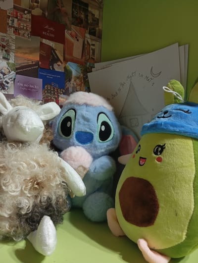 This image shows a close-up of three plush toys arranged on a flat surface, likely a table or desk, against a backdrop of posters and a green wall. The toys are a fluffy white sheep with curly brown and white wool, a blue Stitch-like character with large eyes and a pink ice cream cone, and a green avocado character wearing a blue hat. The background consists of a collage of various posters, some with text and images related to travel, beauty products, and cityscapes. One poster depicts the phrase "Healthy & Wealthy," and another shows a passport. A drawing on a piece of paper is partially visible behind the toys, featuring a house, a moon, and stars, with handwritten text that appears to read "Me Back To The Night We Met is." The lighting suggests an indoor environment, possibly a bedroom or study. The scene does not contain any people or specific identifiable landmarks of Kokand City, Uzbekistan.