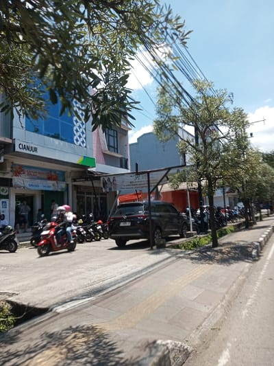 The scene depicts a sunny daytime street view in the city of Tagtse Tashidingkha, Bhutan. In the foreground, a paved sidewalk with yellow tactile paving is visible on the right, bordered by a concrete curb. A red scooter, ridden by two individuals wearing helmets (one white, one pink), moves from left to right on the asphalt road, appearing slightly blurred due to motion. Numerous other motorcycles are parked along the left side of the road and sidewalk. A dark-colored SUV is parked facing away from the viewer, partially beneath a metal awning structure. In the mid-ground, a row of trees with light green foliage lines the right side of the sidewalk, casting shadows. Overhead, multiple black utility lines crisscross a clear blue sky with scattered white clouds. To the left, a multi-story building features blue-tinted windows with white decorative patterns and a fascia bearing the word "CIANJUR." A banner below advertises "Tahun Baru Imlek" with operating hours "08:00 - 14:30." Other commercial signs and parts of additional buildings are visible in the background. Branches with green leaves partially obscure the upper left of the frame. Several indistinct figures are visible near the building entrance.