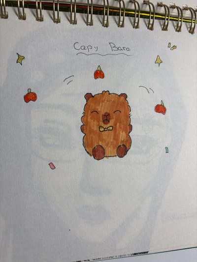 The image shows a handwritten drawing of a brown, fluffy capybara with a yellow bow tie, surrounded by small red fruit shapes, yellow stars, and pink abstract shapes. The text "Capy Bara" is written above the capybara. The drawing is on a white, spiral-bound notebook page. The background of the page has a faint, blurry sketch of what appears to be a human face. There is no information in the image that would allow for a description of the location in the city of Velbert, Germany.