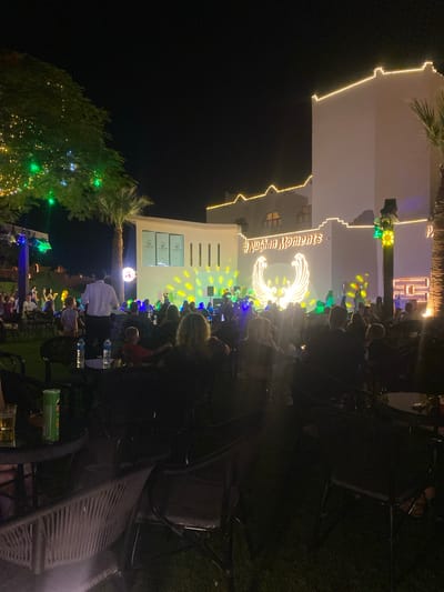 This image captures an outdoor evening event in an unknown location in Egypt. The scene features a building illuminated by decorative lights along its roofline and facade. In front of the building, a stage is set up with lighting effects and a large projection of a winged emblem. A crowd of people is gathered in front of the stage, many standing and some seated at tables in the foreground. A tree decorated with string lights is visible on the left side of the image, and palm trees are also present. The overall atmosphere suggests a celebration or performance.