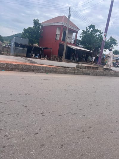The image depicts a street scene in Kona Garu, Nigeria. The perspective is from ground level, looking outwards towards the street and the buildings lining it. A prominent red building with a white-trimmed roof is visible on the left, featuring an open-air section with a balcony. Adjacent to it, several makeshift stalls with corrugated metal roofs line the street, offering various goods.
Several people are present, including a man in a light-colored traditional outfit standing near a purple pole. Other individuals are seen near the stalls, some appearing to be vendors and customers. A few motorcycles are parked in the background, suggesting local transportation. The ground is asphalt, with some debris and small puddles scattered across its surface. The sky is overcast with grey clouds, indicating a potentially cloudy or rainy day. The overall atmosphere appears to be that of a bustling local market or commercial area. There are no discernable emotions conveyed by the people in the image. Some text is visible on posters on the purple pole, but it is too small and blurry to read.