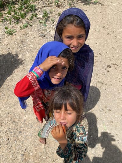 Three young girls stand together on a dusty ground in Ashoqah, Afghanistan. The girl in the back wears a dark blue headscarf and looks directly at the camera with a slight smile. The girl in the middle wears a bright blue headscarf and red patterned shirt, her hand shielding her eyes. The girl in the front has dark hair with bangs, is barefoot, and is holding a piece of food to her mouth. Shadows indicate it is daytime.