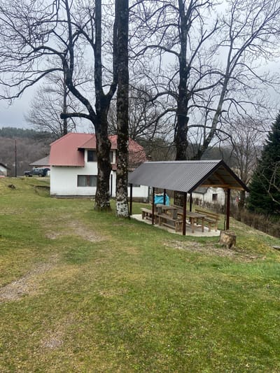 The scene is an outdoor setting in Kolašin, Montenegro. A white building with a pink roof is partially obscured by a bare tree. Nearby is a picnic shelter with a corrugated metal roof and wooden benches. A grassy area extends from the building toward the shelter. The weather appears overcast.