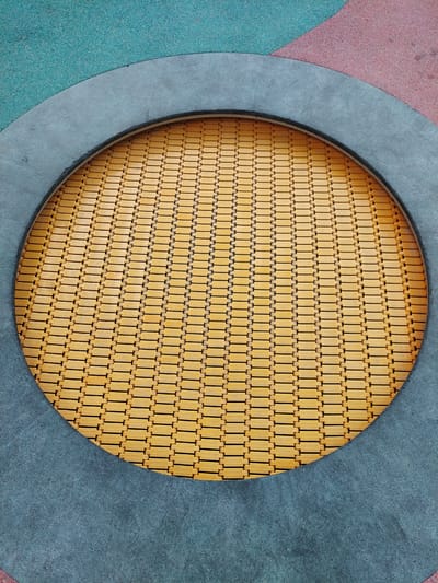 This is a high-angle, close-up shot of a circular trampoline embedded in a playground surface. The trampoline bed is made of interlocking yellow rubber or plastic tiles arranged in a grid pattern. A thick, dark gray border surrounds the trampoline. The playground surface surrounding the gray border is made up of irregularly shaped sections of textured material in teal and pinkish-red. The scene is devoid of people and any other discernible objects. This image likely depicts a public playground area, possibly within the city of Alikonovka, Russia, given the context provided.