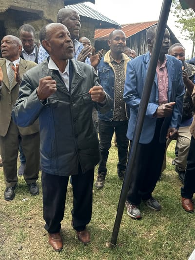 A group of adult and older adult men are gathered outdoors in Kiganjo, Kenya, standing closely together on a grassy patch of ground. The scene captures them in a moment of apparent unity or strong expression. Several individuals, prominently the man in the foreground wearing a dark leather jacket and another man in a blue jacket positioned behind a horizontal dark pole, have their fists clenched. Their expressions are serious and focused, conveying determination or solidarity.

The men are dressed in various casual and semi-formal attire, including denim jackets, a leather jacket, suit jackets, patterned shirts, and trousers. Footwear ranges from dress shoes to sneakers. In the background, a structure with a corrugated metal roof is partially visible on the left, suggesting a semi-urban or rural setting. The ground is covered with green grass interspersed with some patches of bare earth.

The photograph was taken during the daytime, under natural light, and the weather appears fair. No visible text is present in the image. The arrangement and postures of the men suggest they are participating in a gathering or event where shared sentiments are being expressed.