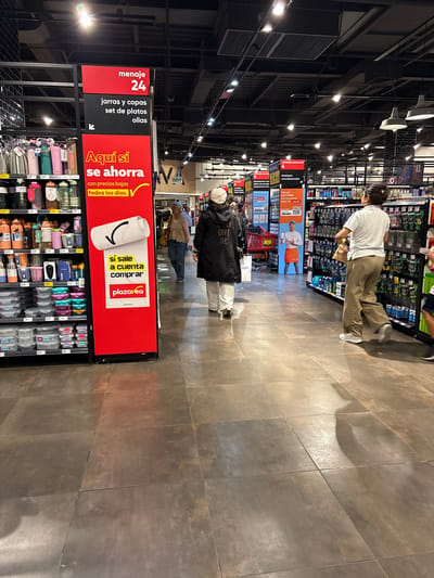 This image captures the interior of a Plaza Vea supermarket in San Isidro, Peru, depicting a wide aisle with various products and shoppers. The scene is brightly illuminated by numerous overhead industrial-style lights against a black ceiling with exposed ventilation ducts. On the left, a tall red and black display sign dominates the foreground. The red top section reads "menaje 24" (housewares 24), followed by a black section listing "jarras y copas" (jugs and glasses), "set de platos" (plate set), and "ollas" (pots), with an arrow pointing left. Below this, a large red section states "Aquí sí se ahorra" (Here you really save) with "con precios bajos todos los días." (with low prices every day.) and a yellow checkmark. A white scroll-like image with a black checkmark is centered below, above a yellow section that reads "sí sale a cuenta comprar" (yes, it's worth buying). The base of the sign displays the "plaza vea" logo with five stars. Adjacent to the sign on the left are multiple shelves stocked with household items. Visible products include a variety of water bottles in different colors and materials, clear plastic food storage containers with colored lids, and other kitchenware. Price tags are visible on the shelves. Three individuals are prominently featured in the aisle. In the immediate foreground on the left, a person, seen from the back, wears a white or light-colored beanie, a dark coat with partial text "GIVE UP" visible on the back, and white pants, carrying a white shopping bag labeled "MARIKI" or similar. Further down the aisle on the left, an older woman with light hair, wearing a light-colored top and blue jeans, pushes a shopping cart with a red basket, looking slightly right. Behind her, a portion of another person in a blue shirt is visible. In the mid-ground on the right, another person, also seen from the back, has dark hair, wears a white polo shirt, loose-fitting tan or khaki pants, and light-colored sneakers. This individual carries a light-colored bag in their right hand and appears to be walking purposefully along the aisle. Farther to the right and partially obscured, a child's legs and arm are visible, wearing blue shorts. The right side of the aisle features additional shelving units displaying various products, likely personal care items, as indicated by signs in the background reading "cuidado personal" (personal care), "shampoo," and "acondicionador" (conditioner). A digital display on