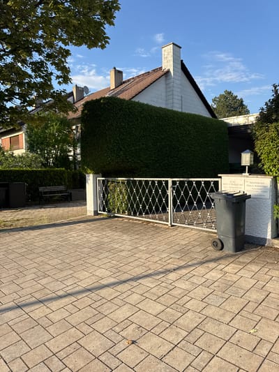 The image shows the exterior of a building in Höhenkirchen-Siegertsbrunn, Germany. The building appears to be a clinic or practice, as indicated by the signage. The text on the sign reads: "Naturheilpraxis Petra A. Kratschmann Heilpraktikerin" and "gemeind Walter A. Kratschmann Facharzt für Allgemeinmedizin". This translates to "Naturopathic practice Petra A. Kratschmann Heilpraktikerin" and "community Walter A. Kratschmann Specialist in general medicine".

The scene is an outdoor shot, likely taken during the day, as suggested by the bright sunlight casting shadows. The weather appears clear with a blue sky and some wispy clouds. The foreground is paved with interlocking bricks, and a grey garbage bin stands next to a white metal gate. Behind the gate, a well-maintained hedge forms a screen. A portion of a white building with a brown roof and a chimney is visible. Trees with green foliage are also present in the scene, suggesting a garden or natural surroundings. The overall impression is one of a clean and orderly environment, possibly a professional practice with a welcoming outdoor space. There are no people visible in the image.