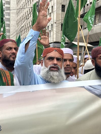 In this image, several men are gathered, with one man in the foreground prominently featured. He has a gray beard and is wearing a light blue shirt and a reddish-brown skullcap. His right hand is raised, palm facing outwards, as if addressing a crowd. Behind him, other men are visible, also wearing skullcaps and traditional attire. Some are looking towards the foreground, while others are looking elsewhere.
Green flags, some with white text or symbols, are visible in the background, held aloft on bamboo poles. These flags suggest a gathering or rally. The setting appears to be outdoors, possibly in an urban environment, with a building structure visible in the background, featuring a grid-like pattern. The lighting suggests it is daytime, and the atmosphere seems to be one of engagement or demonstration. The man in the foreground's expression and raised hand convey a sense of purpose or communication. The overall scene captures a moment during a public gathering in Karachi Division, Pakistan.
