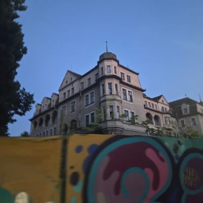 The image shows a large, old building with a light-colored facade, likely a former hotel or sanatorium, set against a clear blue sky. The building has multiple stories, ornate architectural details, pitched roofs, and several windows, some of which appear broken. A prominent turret with a pointed roof stands out on one corner. Trees are visible on the left side of the image, and a section of a wall covered in colorful graffiti is in the foreground, obscuring the lower part of the building. The overall impression is one of a grand but possibly abandoned or neglected structure.

This building is identified as the Royal Palace (Kráľovský palác), located in the spa town of Piešťany, Slovakia. It was built in 1904-1905 by the architect Koloman Kludovanský, and it served as a sanatorium and hotel.