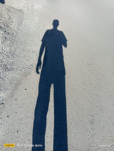In the city of Kurgantepa, Uzbekistan, a shadow of a person stands on an asphalt surface. The shadow is long and stretches across the frame, indicating it was captured during daylight hours. The ground appears to be a mixture of asphalt and small stones, suggesting a road or pathway. The lighting indicates a sunny day. The image contains a watermark in the bottom left corner that reads "realme Shot by бек фото" and in the bottom right corner "realme 8i".