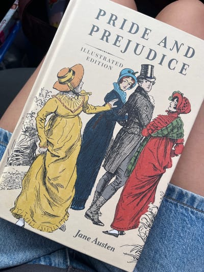 The image displays an off-white hardbound copy of "Pride and Prejudice Illustrated Edition" by Jane Austen, held open-faced on the lap of an unidentified individual. The book's cover features a colorful illustration depicting four Regency-era figures: from left, a woman in a yellow dress and straw hat; a woman in a dark blue coat, light blue ruffled collar, and blue bonnet; a man in a black top hat, dark coat, and light grey breeches; and a woman in a red dress, patterned green and red shawl, and red bonnet. The figures are depicted in a staggered line, with the woman in blue extending her hand towards the man, who gazes forward. The book rests against the person's bare upper thighs, with light blue denim fabric visible below and to the right of the book. The background includes dark upholstery or fabric in the upper left, alongside a blue plastic container with a partially visible white label (text beginning "Che..."). No discernible visual information within the image indicates the specific location within the city of El Paso, United States.