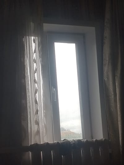 A vertical, narrow window with a white frame is centered in the frame, partially obscured by sheer, patterned curtains on either side. The curtains on the left are lighter and more translucent, with a subtle leaf-like pattern, while the curtain on the right is darker and more opaque, with a more prominent floral design. The window pane itself is bright white, suggesting an overcast or very bright sky outside. Below the window is a dark, ribbed radiator, its top edge softened by a draped fabric or material, possibly another curtain or a blanket. The overall lighting is dim, creating a contrast between the bright window and the darker foreground. The scene appears to be an interior view of a room. There are no people or animals visible. The location is specified as Lobanovo, Kazakhstan.