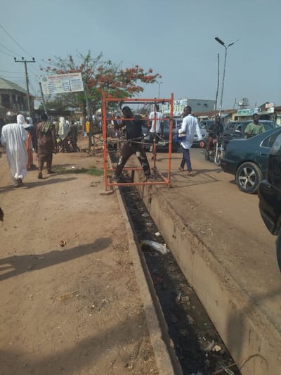 The scene is a street in New Karu, Nigeria, located at latitude 8.985 and longitude 7.565. A man in a black shirt and brown pants is standing in a red metal framework next to a concrete drainage ditch. He is facing the camera, and his arms are extended to the side as if he is stretching. There are several other people in the background, walking on the sidewalk or driving motorcycles. The ditch is filled with garbage and debris, including plastic bags, bottles, and food wrappers. The street is dusty and there are some trees in the background. The sky is clear and blue. The buildings are tall and white, and there are some signs in the background. The scene suggests a typical urban environment in Africa. The image shows the state of the drainage system in the area.